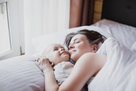 Happy young mother and her baby playing in bed with white linens. Happy family concept.の写真素材