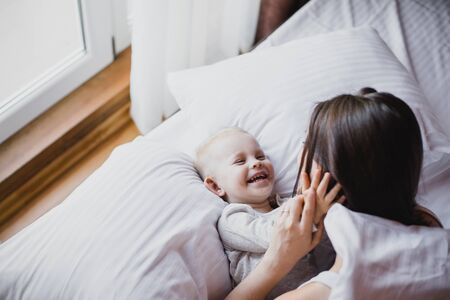 Happy young mother and her baby playing in bed with white linens. Happy family concept.