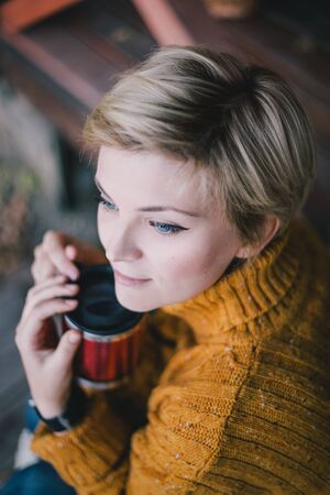 Beautiful woman blond with short haircut and blue eyes sitting near lake with cup of hot tea. Cozy fall. Slow living concept.の写真素材