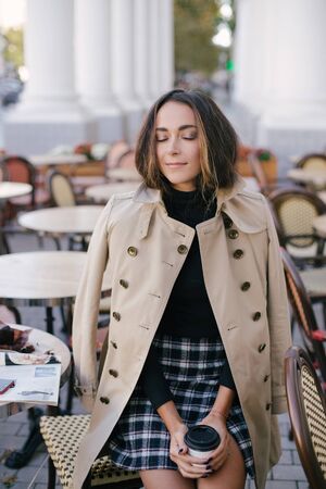 Young beautiful woman in trendy trench coat  drinking coffee in a street cafe.の写真素材