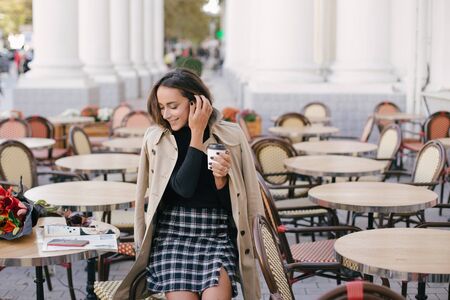 Young beautiful woman in trendy trench coat  drinking coffee in a street cafe.の写真素材
