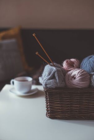 Balls of woolen yarn and knitting needles in basket and hot herbal tea in cozy interior.の写真素材