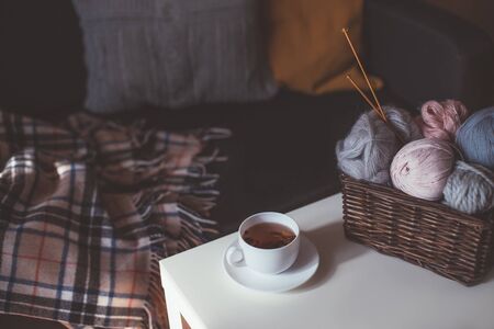 Balls of woolen yarn and knitting needles in basket and hot herbal tea in cozy interior.