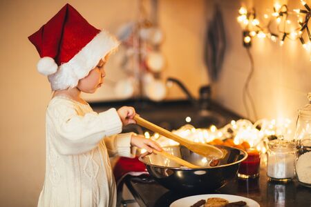 Cute baby girl in Santa Claus hat making Christmad cookies on decorated kitchenの写真素材