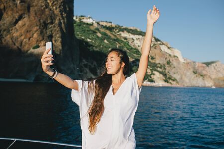 Beautiful young woman taking selfie on her smart phone on yacht with ocean view on background.の写真素材