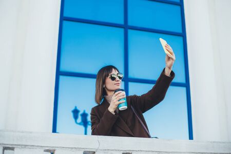 Young beautiful stylish woman drinking coffee and using her smart phone on city street.の写真素材