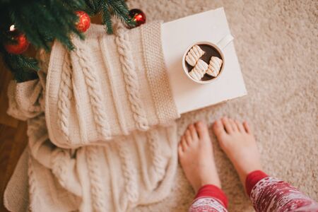 Hot cocoa with marshmallows and woman feet in holiday pajamas near Christmas tree with festive lights on the background.の写真素材