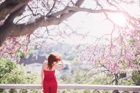 Young woman in red jumpsuit posing near blooming tree in spring city.