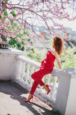 Young woman in red jumpsuit posing near blooming tree in spring city.