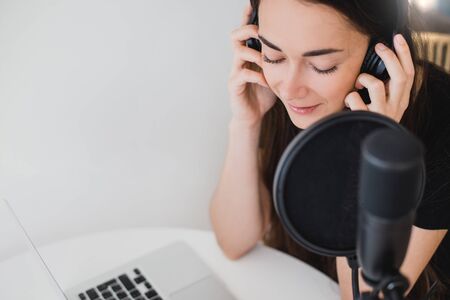 Attractive young woman blogger with long black hair recording online podcast using her laptop, headphones and professional microphone.の写真素材