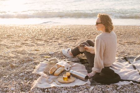 Young woman in knitted sweater having picnic at beach with tea in flask and fresh baguettes in wicker eco bag. Cozy slow lifestyle concept.の写真素材