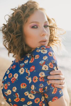 Young beautiful stylish woman with curly messy hair wearing blue dress posing at beach on sunset time.の写真素材