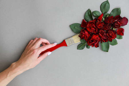 Creative layout with paint brush and red rose flowers on gray background. Woman hand hold brush paints. Copy spaceの写真素材