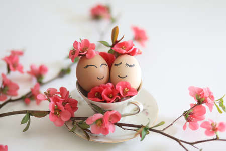 Creative easter eggs with cute face, sleepy eyes and spring flowers in tea cup on white background....