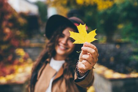 A beautiful smiling curly blond long-haired young girl in a brown hat with wide brim and a beige coat shows yellow maple leaf in frame. Focus on the yellow maple leaf.の写真素材