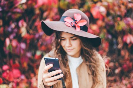 Beautiful curly young girl in a brown hat with wide brim and a beige coat looks at the phone against the background of an autumn red, raspberry vineyard in the afternoon. Focus on the phone.の写真素材