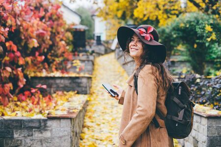 Curly young girl in a brown hat and a beige coat walks in the autumn park with a phone and a...