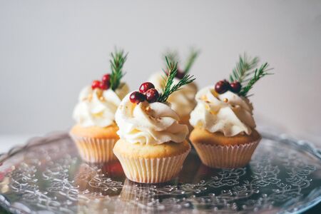 Beautiful christmas cupcakes with curd cream, cranberries and spruce branches on top on the glass table against a white wall.の写真素材