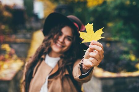 A beautiful smiling curly young girl in a brown hat with wide brim and a beige coat shows yellow maple leaf in frame. Stands in the autumn park. Focus on the yellow maple leaf.の写真素材