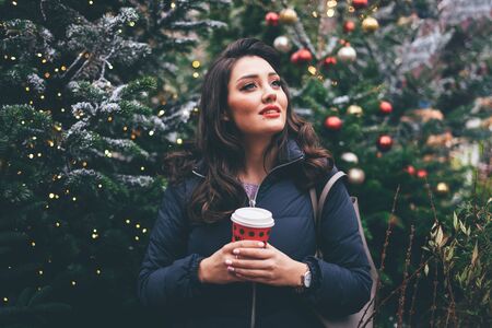 Beautiful young woman with makeup in winter clothes with red paper cup standing near Christmas trees...