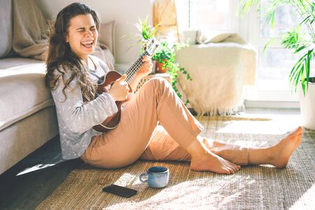 Young beautiful woman plays ukulele, sings and laughs. Sitting at home on the floor in the sun.の写真素材