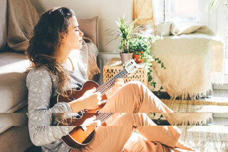 Young beautiful woman plays ukulele, sings and looking away. Sitting at home on the floor in the sun.の写真素材