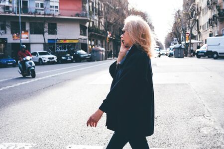 Adult blonde lady with glasses and a dark coat, talking on the phone and cross the street. In the sunset light in Barcelona.の写真素材
