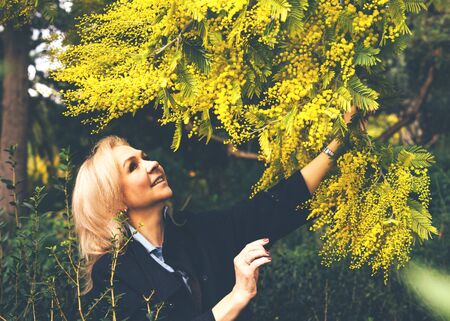 An adult blonde lady in a dark coat looks up at a flowering mimosa tree and smiles. Side view.の写真素材