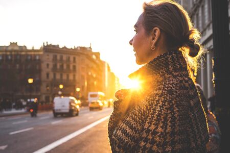 An adult beautiful lady in a plaid coat in the rays of the setting sun stands along the street of barcelona and looks away. View assembly close.の写真素材