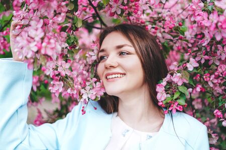 Young brunette girl with a flower behind her ear looks away at a blossoming pink apple tree and smiles. Close up. Holding a flowering branch.の写真素材