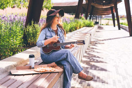 Young pretty woman in hat playing her ukulele outdoors in the modern park and smiling. Carefree mind, concept musical playing, creative activity, music education.の写真素材