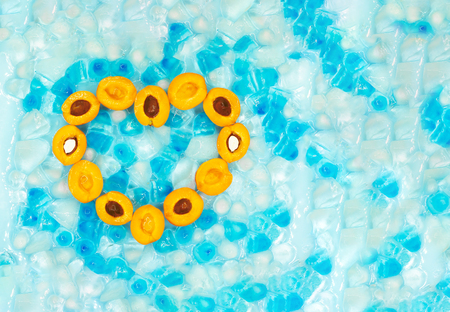 Heart made of ripe apricots with or without stones against the background of transparent and blue ice cubes with copy space. Top view. Fresh fruits. Summer concept.の写真素材