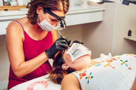 Beautician in beauty center performing an eyebrow treatment to client, with masks during the new...
