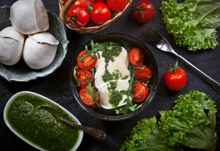 preparing fresh mozzarella salad with lettuce and arugula leaves, pesto sauce, fresh cherry tomatoes and cedar nuts, on a black plate on dark slate table background, nobodyの写真素材