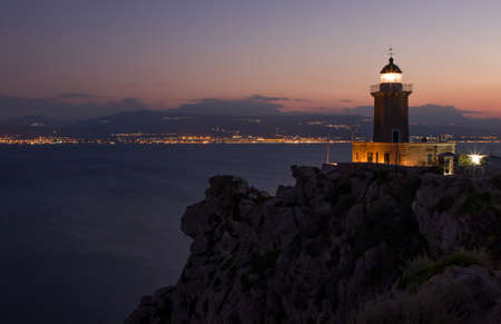 Night landscape, lighthouse on the rocky mountain on the background of night city, Greeceの写真素材