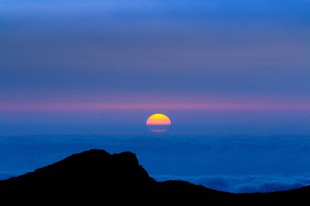 The sun rises between the silhouette of some mountains and a sea of fogの写真素材