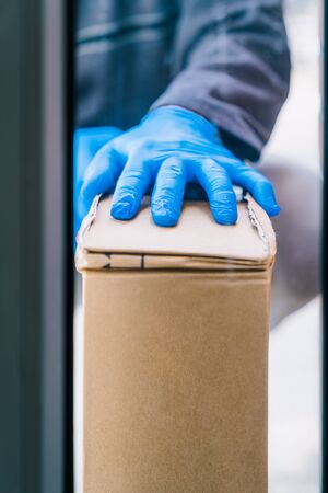Deliverer leaving a package at the customer's door as a measure of protection against the coronavirus. Delivery man wearing blue latex gloves. Delivery man through the glass door.の写真素材