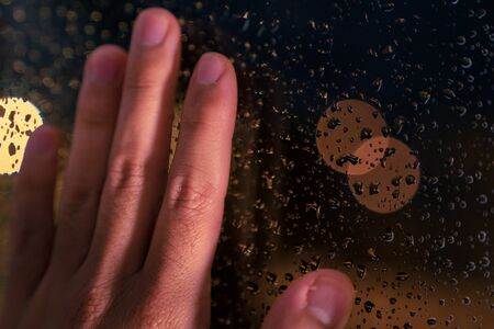 Hand resting on the window at night with the warm light of the street lamps and with drops of water...