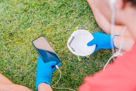Person who plays sports sitting on the grass in a park listening to music with a mobile phone with a mask and blue latex gloves because of the preventive measures needed for the coronavirus.の写真素材