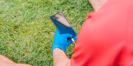 Person who plays sports sitting on the grass in a park listening to music with a mobile phone wearing blue latex gloves because of the prevention measures needed for the coronavirus.の写真素材