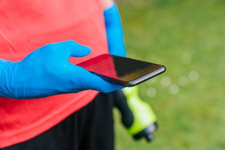 Sportsman consulting the mobile phone in the park with the protection measures to prevent the coronavirus. Boy with blue latex gloves, a bottle of sports water and a cell phone.の写真素材