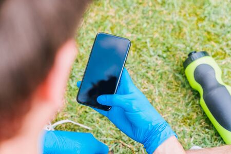 Boy sitting on the green grass of the park consulting his mobile phone with blue latex gloves for the prevention measures needed for the coronavirus.の写真素材