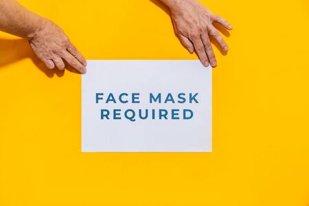 Face mask required. Women's hands hanging a sign for people to respect social distancing measures with an orange background.の写真素材