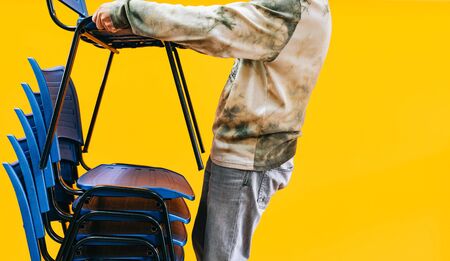 Man picking up blue chairs one on top of another with an orange background. Adaptation of school and academy chairs to the new normal.の写真素材