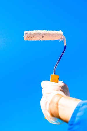 Hand holding a roller of white paint with blue sky in the background. Concept of painting the house.