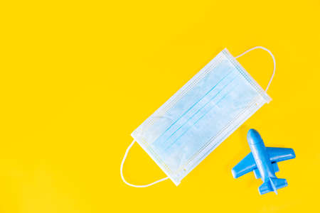Medical face mask and blue toy plane isolated on yellow background. Travel safely on holiday flights to prevent the spread of the virus. Going vacations in summer during the coronavirus pandemic.の写真素材