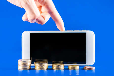 Hand holding a mobile phone with stack of coins on blue background. Business growth and financial saving concept of money. Cell phone with black screen for copy space.の写真素材
