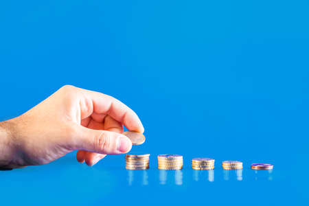 Hand depositing a euro coin in a stack of coins on blue background. Business growth and financial saving of money.の写真素材