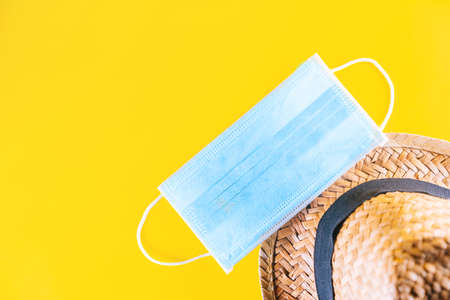 New tourism trend after Covid-19 pandemic. Medical face mask and straw hat isolated on yellow background. Concept of going on holiday in summer while during the coronavirus pandemic.の写真素材