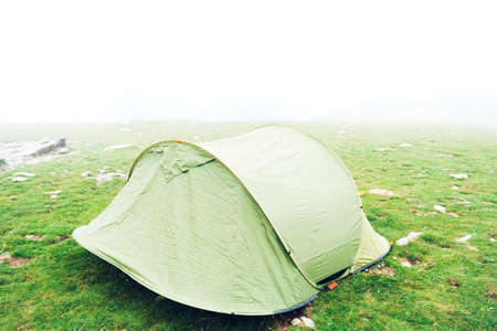 Tent in the wilderness. A single green camping tent in a grassy meadow with fog on the mountain. Trekking, camping, hiking and wild life concept. Real Outdoor Adventure.の写真素材